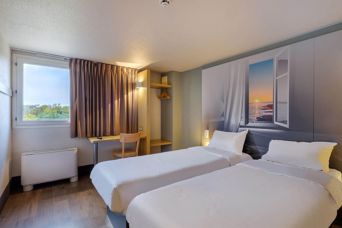 Une chambre d’hôtel avec deux lits, un bureau, une chaise, un radiateur et un tableau représentant un coucher de soleil sur l’océan.