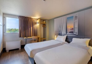 Une chambre d’hôtel avec deux lits, un bureau, une chaise, un radiateur et un tableau représentant un coucher de soleil sur l’océan.