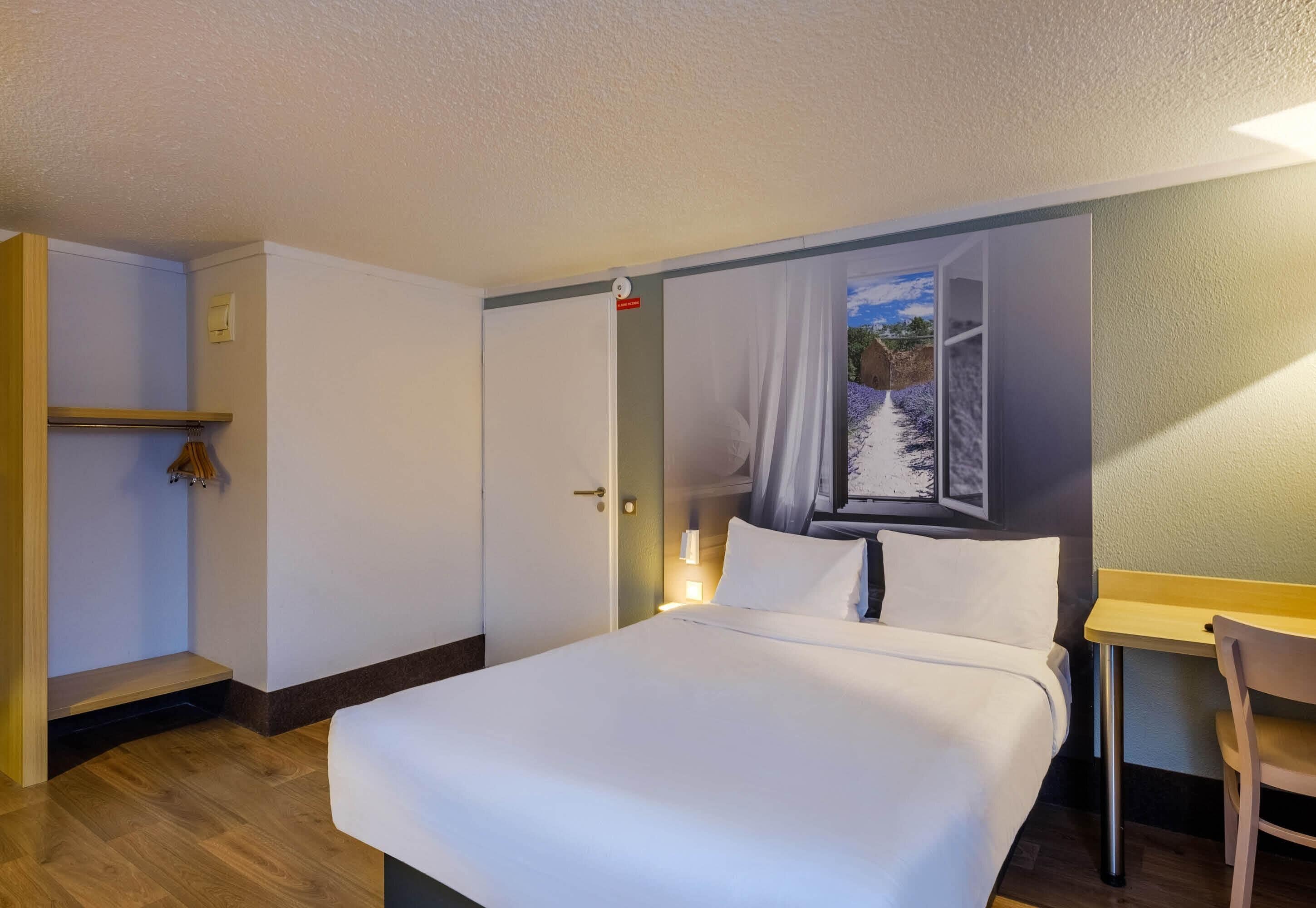 Una habitación de hotel con cama, escritorio, silla, estante y puerta.