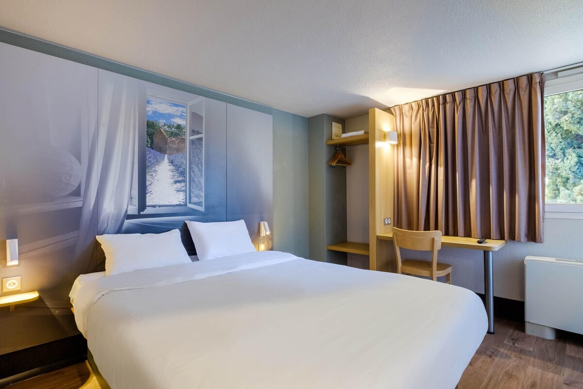 A hotel room with a large bed, a desk, a chair, a window with curtains, and a view of a rural landscape.