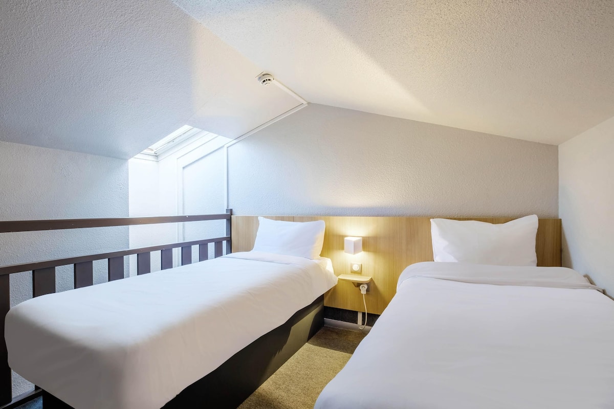 Une chambre d’hôtel avec deux lits, une table de chevet avec une lampe, une fenêtre avec des stores et un luminaire au plafond.