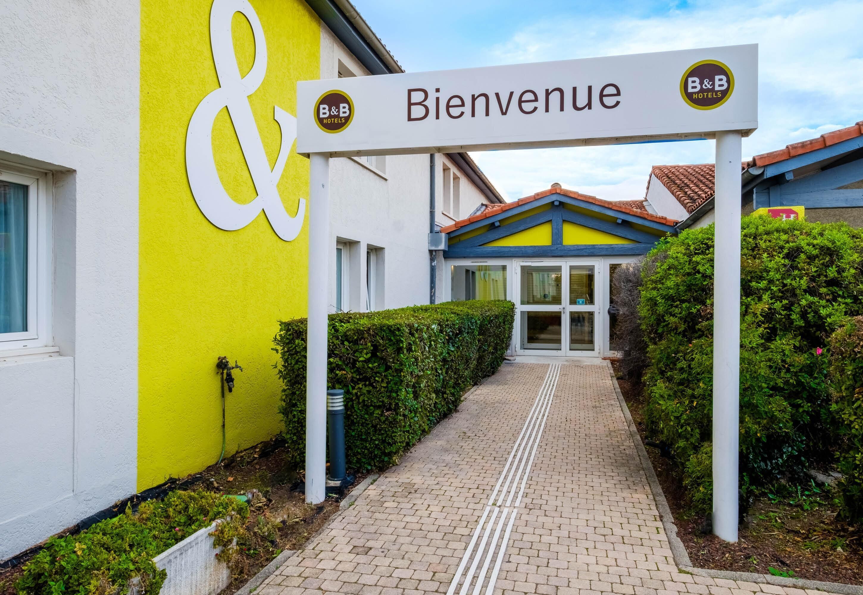 A hotel entrance with a 'Bienvenue' sign, a yellow wall, and a pathway leading to the building.
