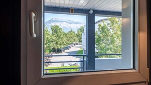 Une fenêtre donnant sur un parking, des arbres et, au loin, une montagne.