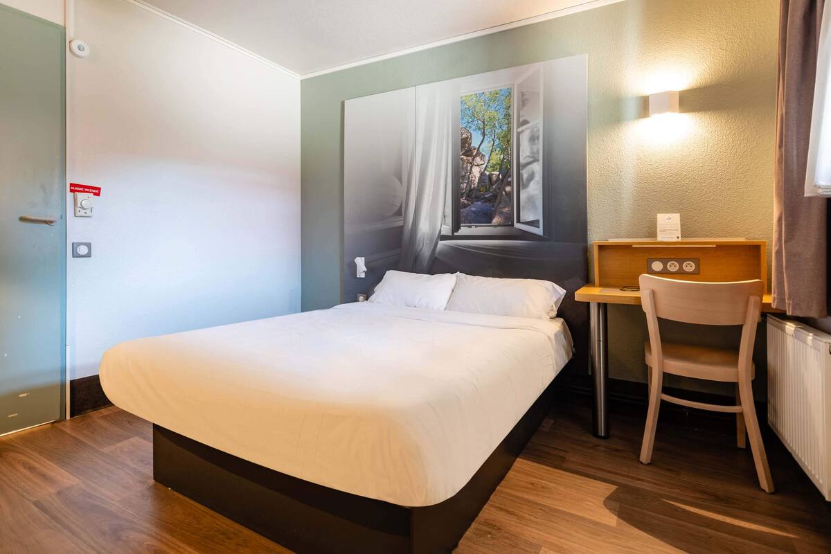 A neatly made bed with a wooden headboard, a small desk with a chair, and a painting of a tree on the wall.