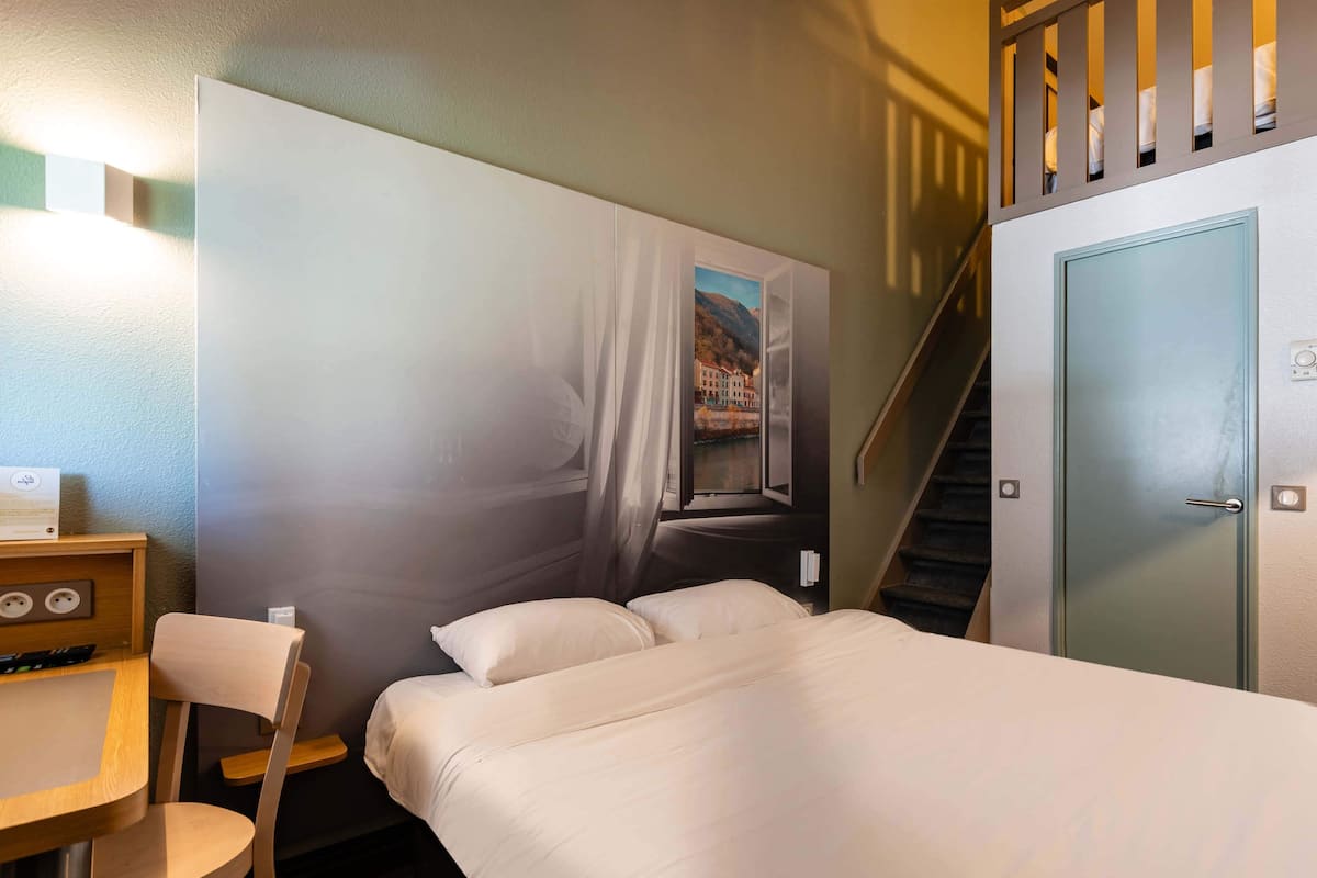 A hotel room with a bed, a desk with a chair, a painting on the wall, and a staircase.