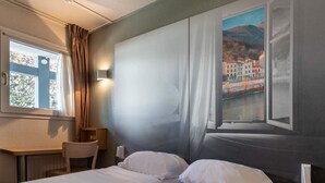 Une chambre d’hôtel avec un lit, un bureau et une chaise. Il y a une fenêtre avec des rideaux, et de là, on peut voir des immeubles et une montagne.