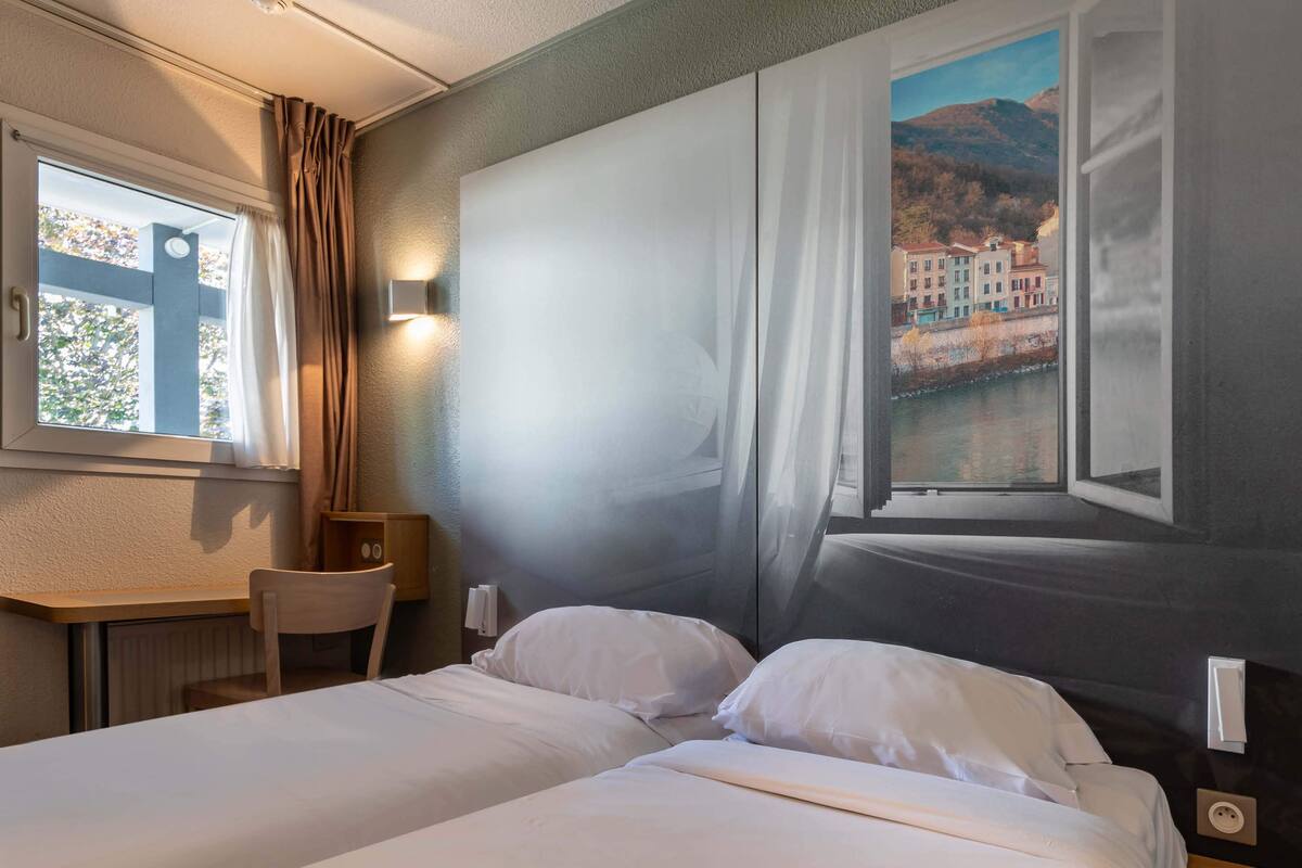 A hotel room with a bed, a desk, and a chair. There is a window with curtains and a view of buildings and a mountain.