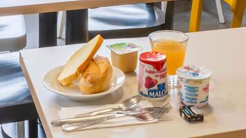 A breakfast table with a plate of toast, a glass of orange juice, a container of yogurt, and a small jar of jam.