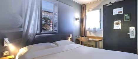 A hotel room with a bed, a desk, a chair, a window with curtains, and a door with a calendar and a sign.