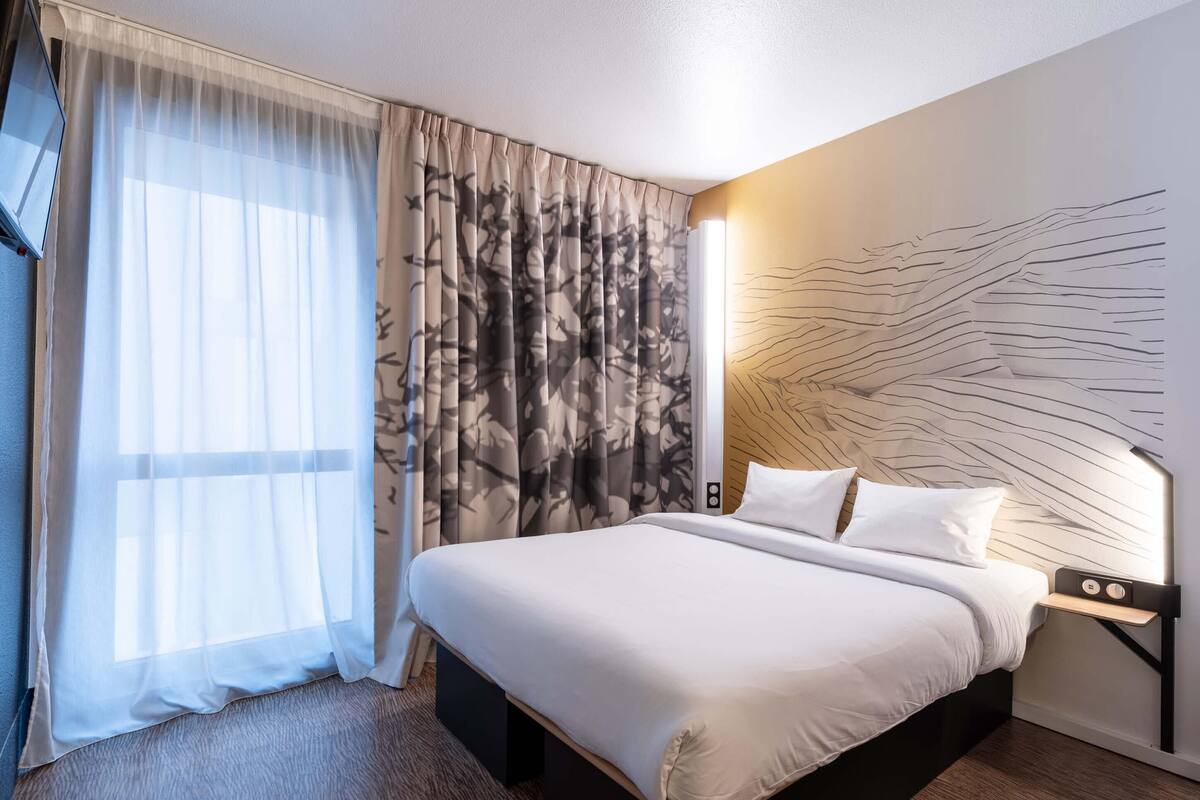 A hotel room with a large bed, sheer curtains, a patterned curtain, a wall art design, and a bedside table with a lamp.