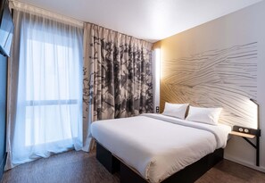 A hotel room with a large bed, sheer curtains, a patterned curtain, a wall art design, and a bedside table with a lamp. - B&B HOTEL Lille Villeneuve d'Ascq Grand Stade 3 étoiles (Villeneuve-d'Ascq)