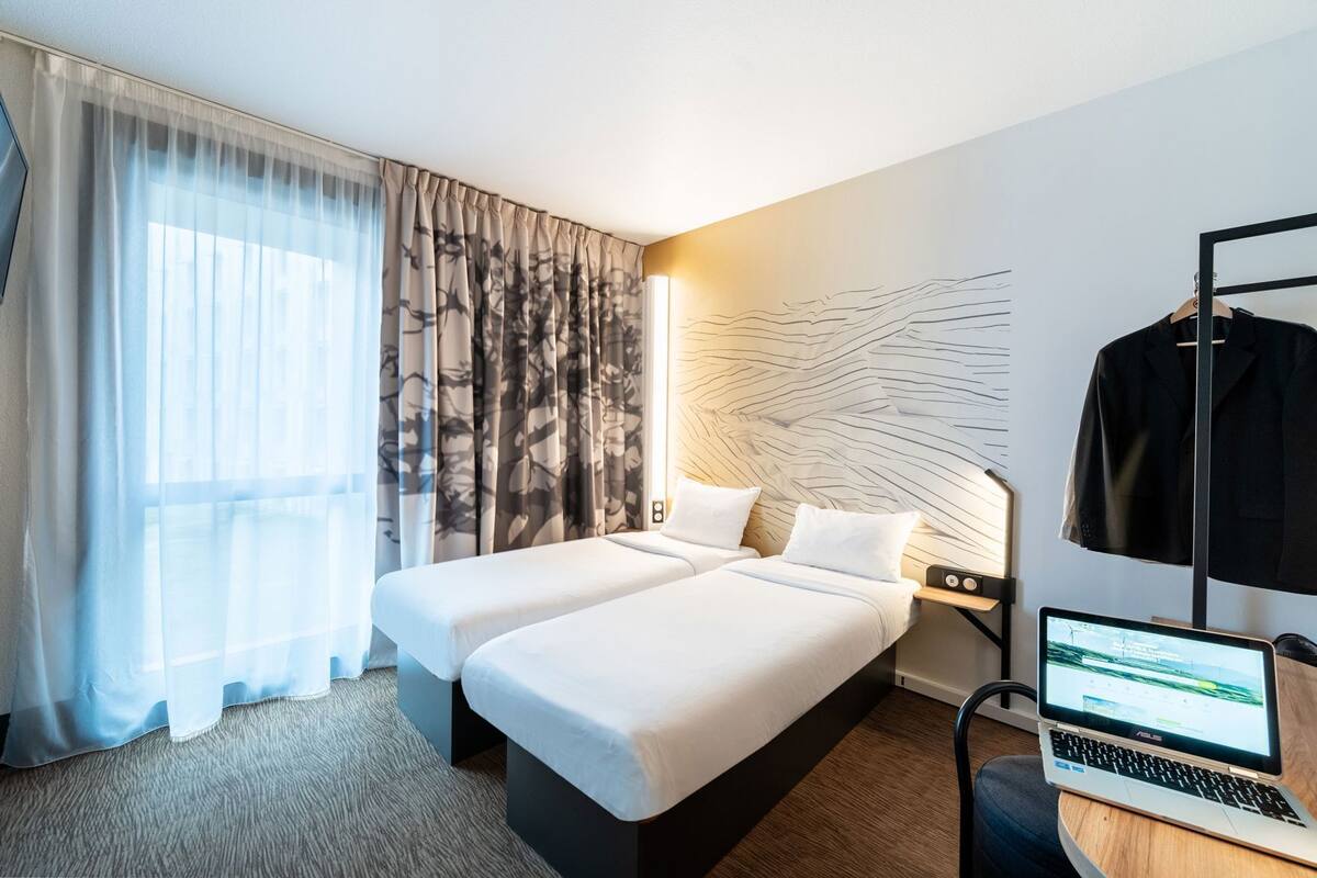 A hotel room with two beds, a desk with a laptop, a coat rack, and a wall with a leaf pattern.