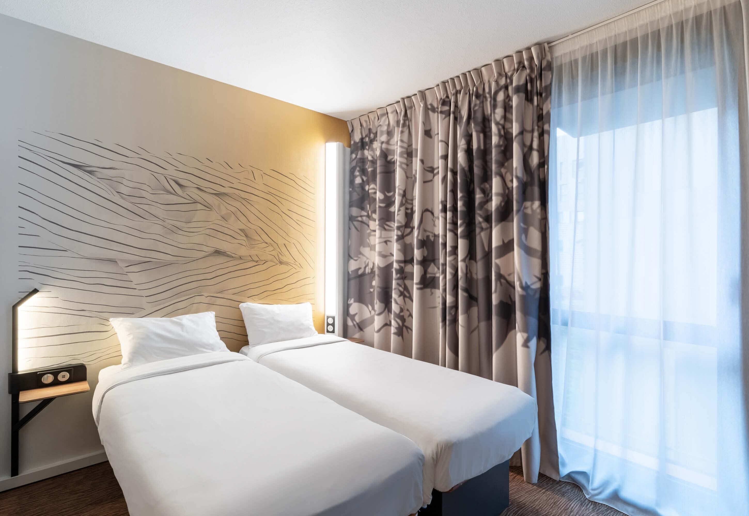 A hotel room with two beds, a wall art piece, a nightstand with a lamp, and sheer curtains.