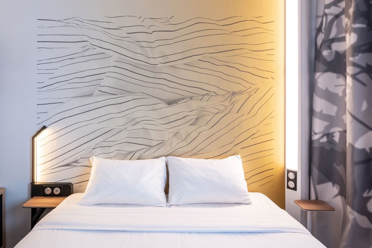 Habitación de hotel con una cama, almohadas blancas, una pared con un diseño ondulado, una mesita de noche con una radio y una ventana con cortina.