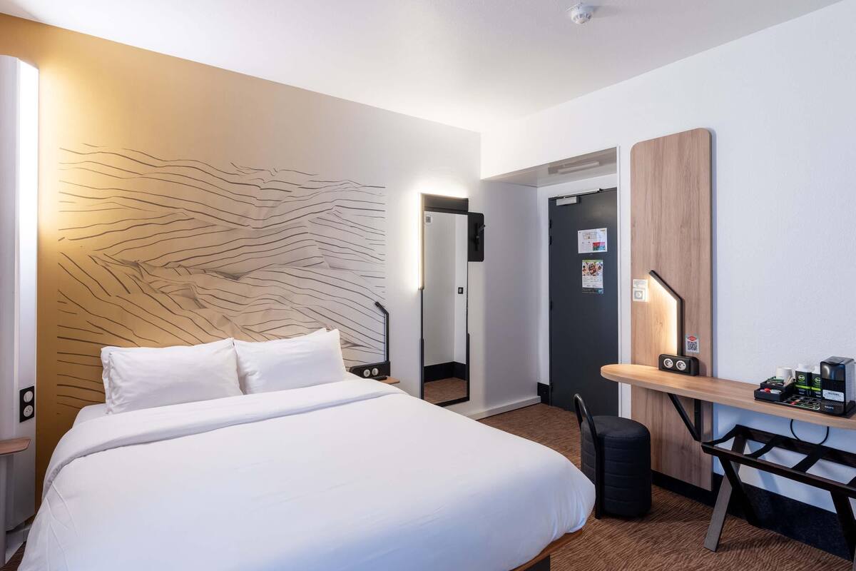A hotel room with a bed, bedside table, desk, chair, and a wall with a unique design.