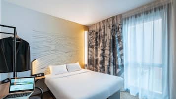 A hotel room with a bed, a desk with a laptop, a coat rack, and a wall with a geometric pattern.