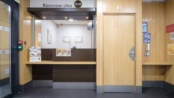 Een receptiebalie met een bord waarop staat: “Bienvenue chez B&B Hotels”. Er hangen ingelijste certificaten aan de muur, een beveiligingscamera en een telefoon.