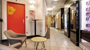 A hotel lobby with a red door, seating area, vending machines, and a corridor leading to other rooms. - B&B HOTEL LILLE Tourcoing Centre (Tourcoing)