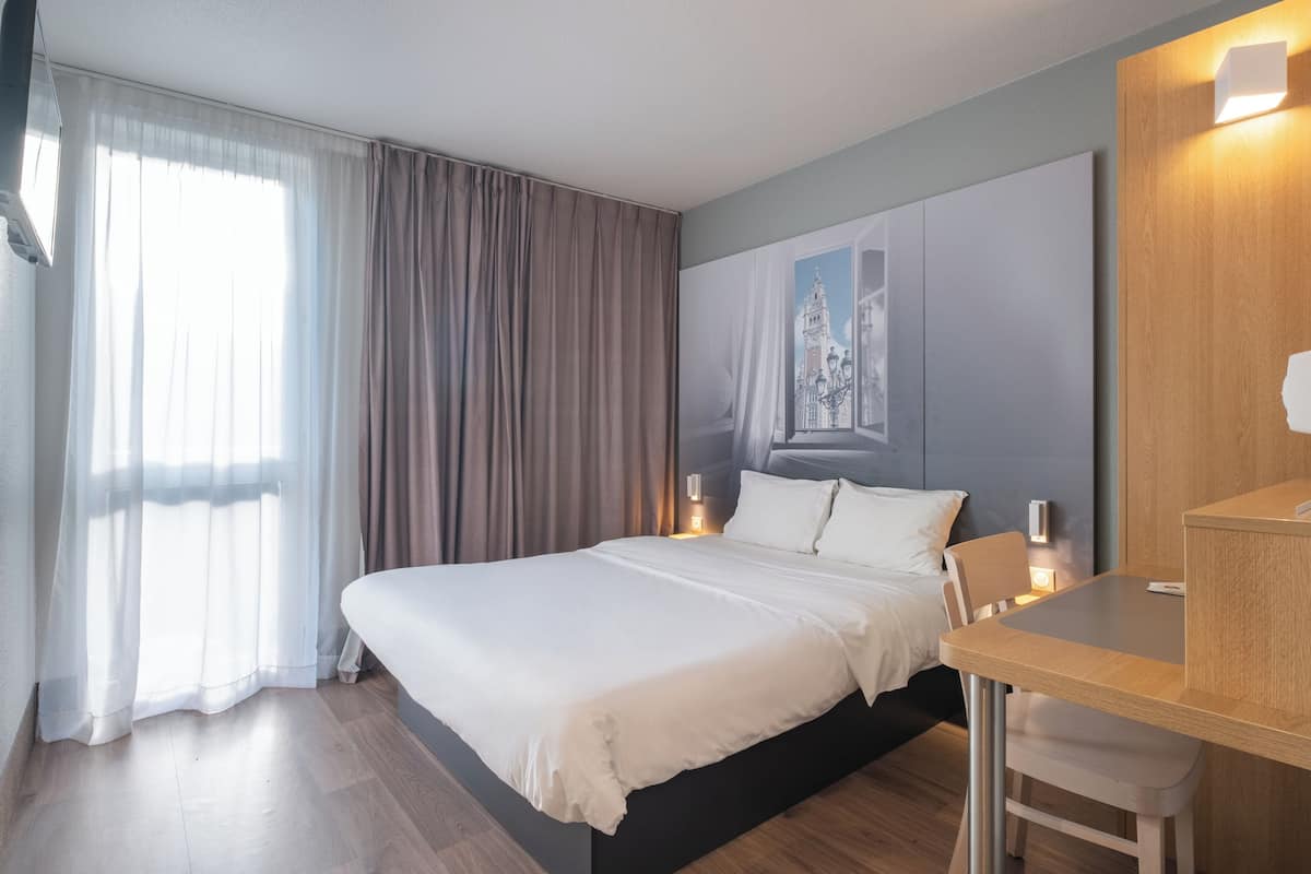 Una habitación de hotel con cama, mesita de noche, silla y un ventanal con cortinas.