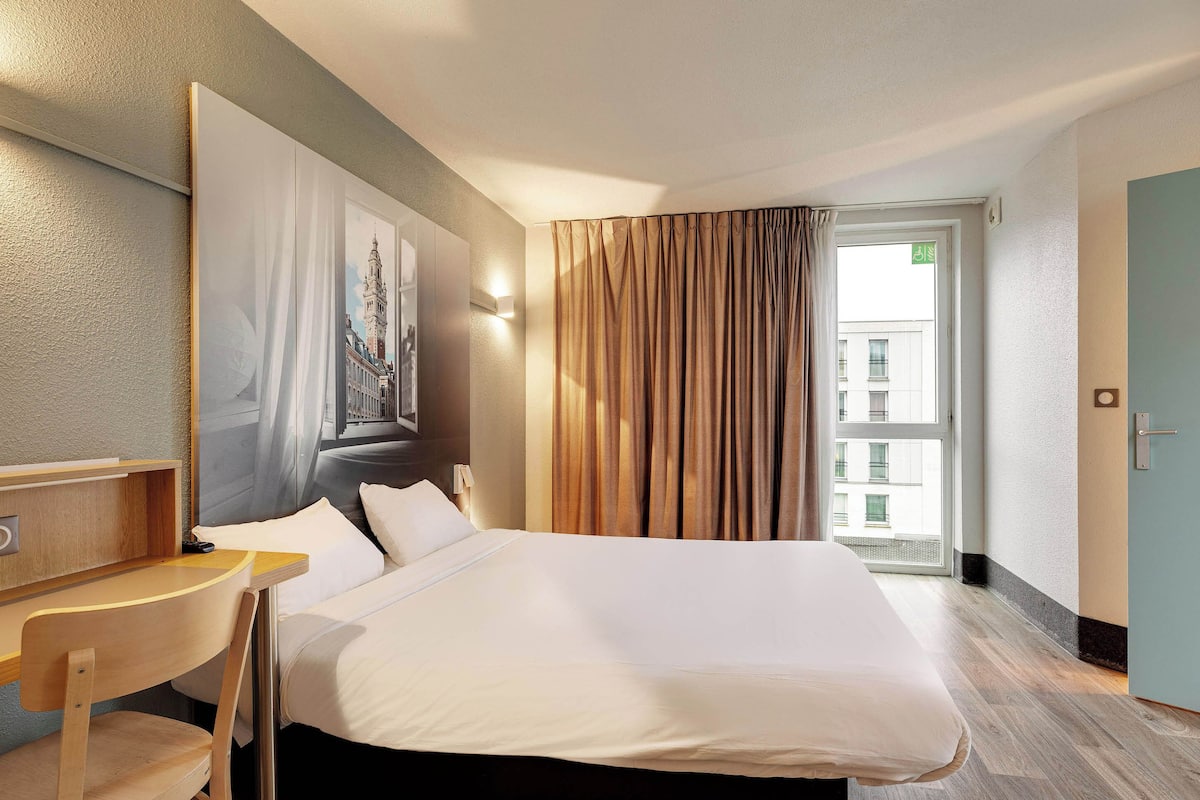 Habitación de hotel con una cama grande, un escritorio, una silla, un espejo y una ventana con cortinas.