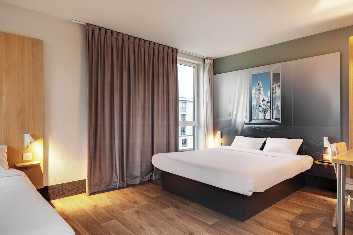 Habitación de hotel con una cama grande, un escritorio, una silla, una mesita de noche y un ventanal con cortinas.