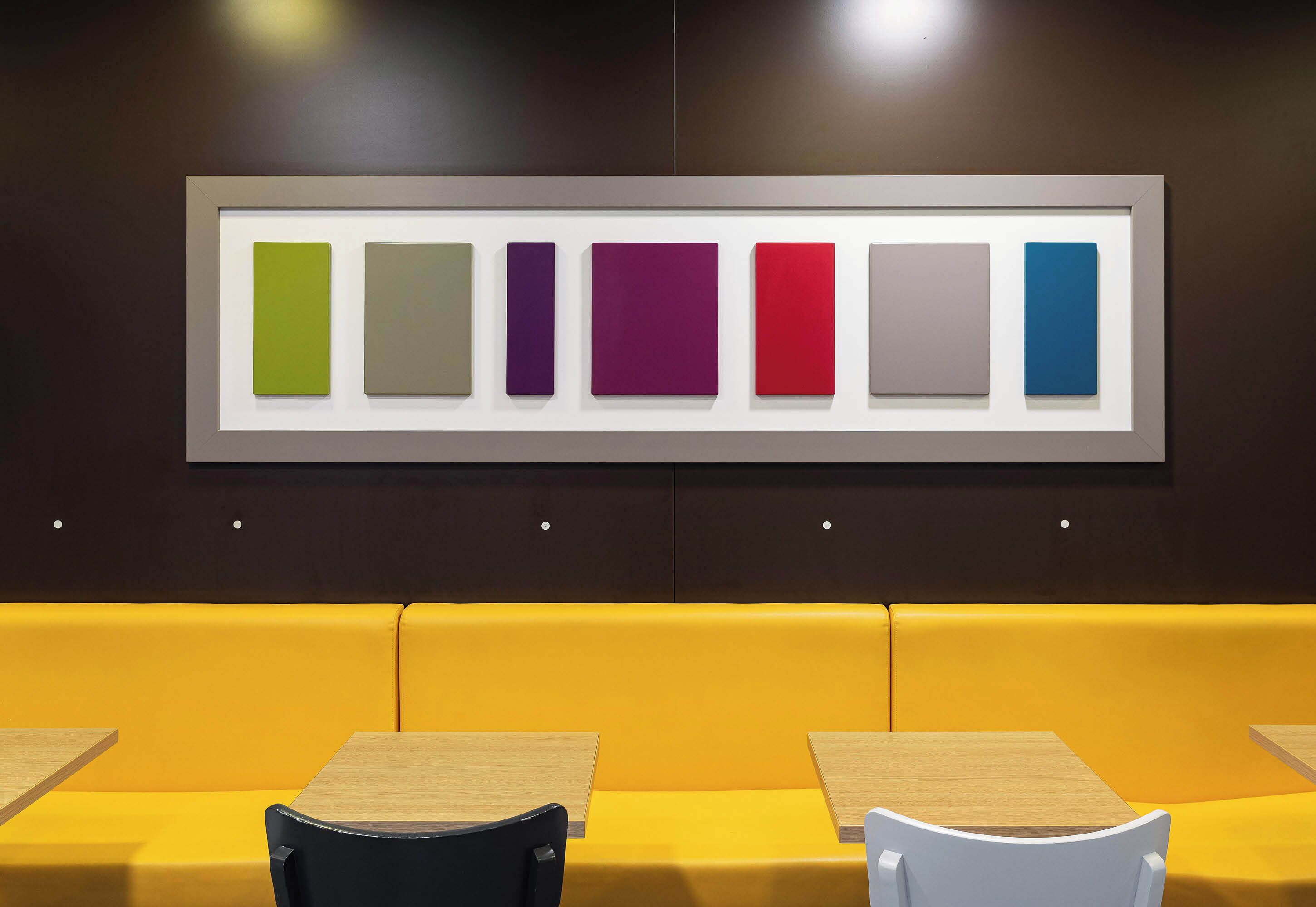 A modern interior with a row of colorful square panels on the wall, a yellow cushioned bench, and wooden tables.