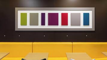 A modern interior with a row of colorful square panels on the wall, a yellow cushioned bench, and wooden tables.