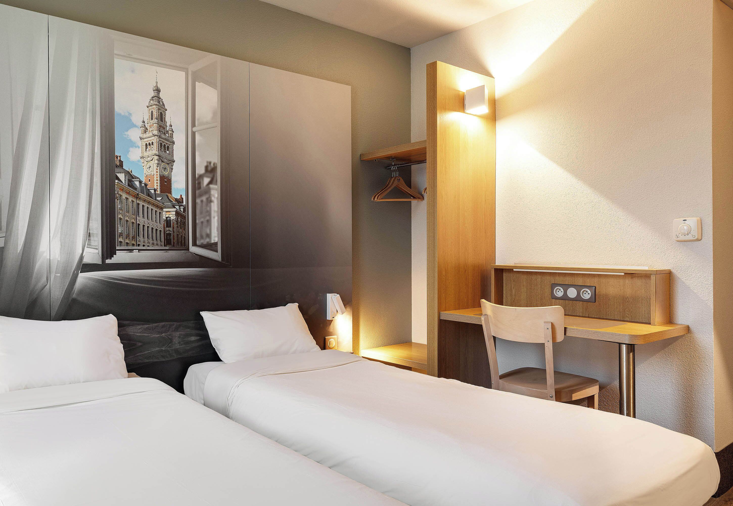 Une chambre d’hôtel avec deux lits, un bureau, une chaise, un porte-manteau et un tableau représentant un paysage urbain.