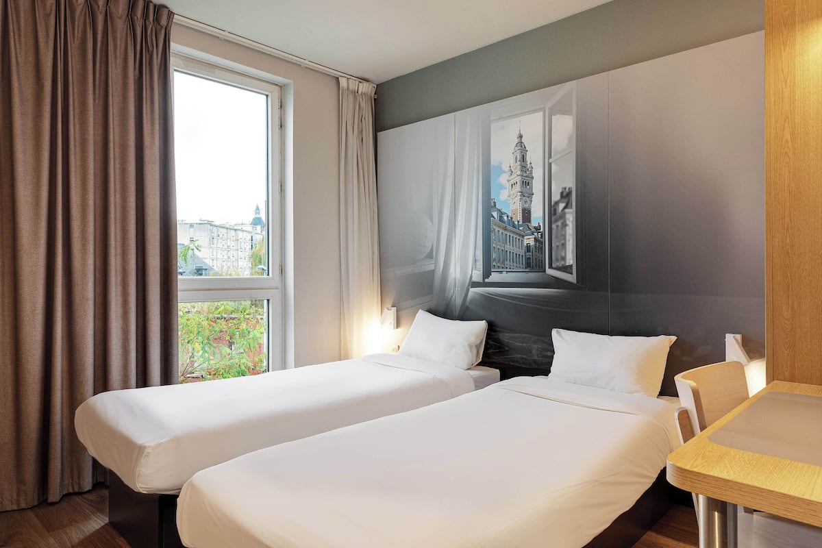 Habitación de hotel con dos camas individuales, un ventanal con cortinas, un cuadro de un edificio y una mesa de madera.