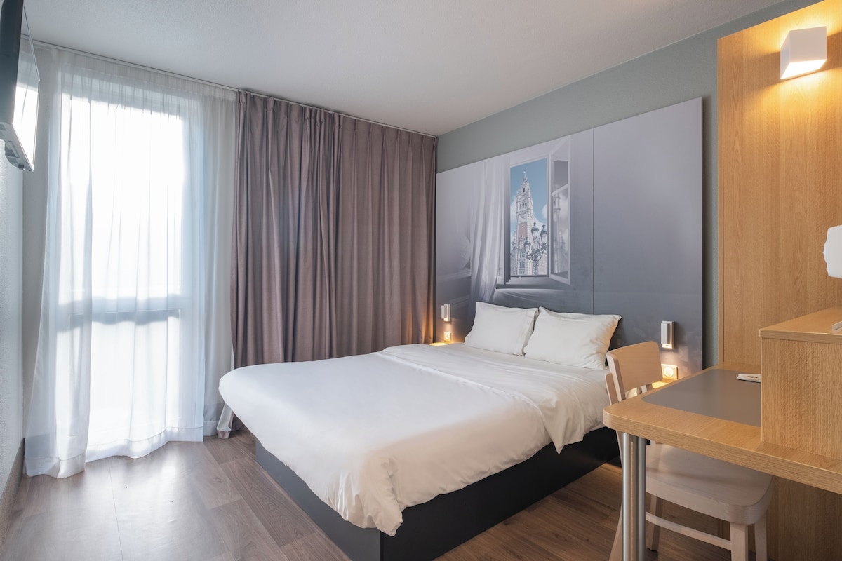 A hotel room with a large bed, a wooden desk, and a nightstand with a lamp.