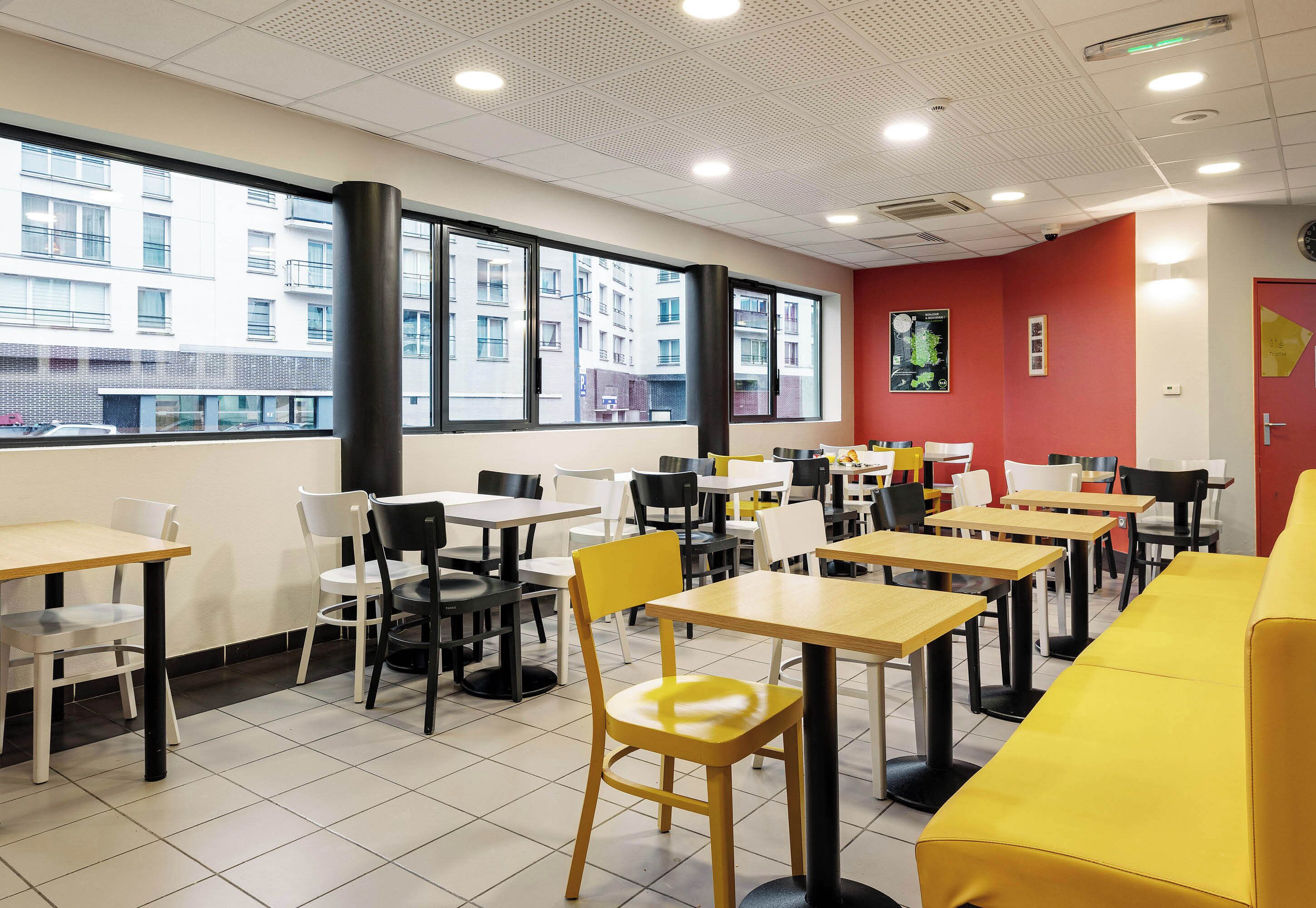 Un comedor moderno con varias mesas y sillas, ventanas amplias con vista a edificios, y una pared decorada en rojo.