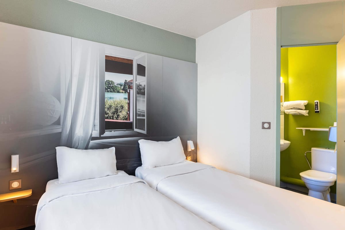 Une chambre d’hôtel avec deux lits, une fenêtre avec des rideaux, un mur vert, un lavabo blanc et des toilettes.