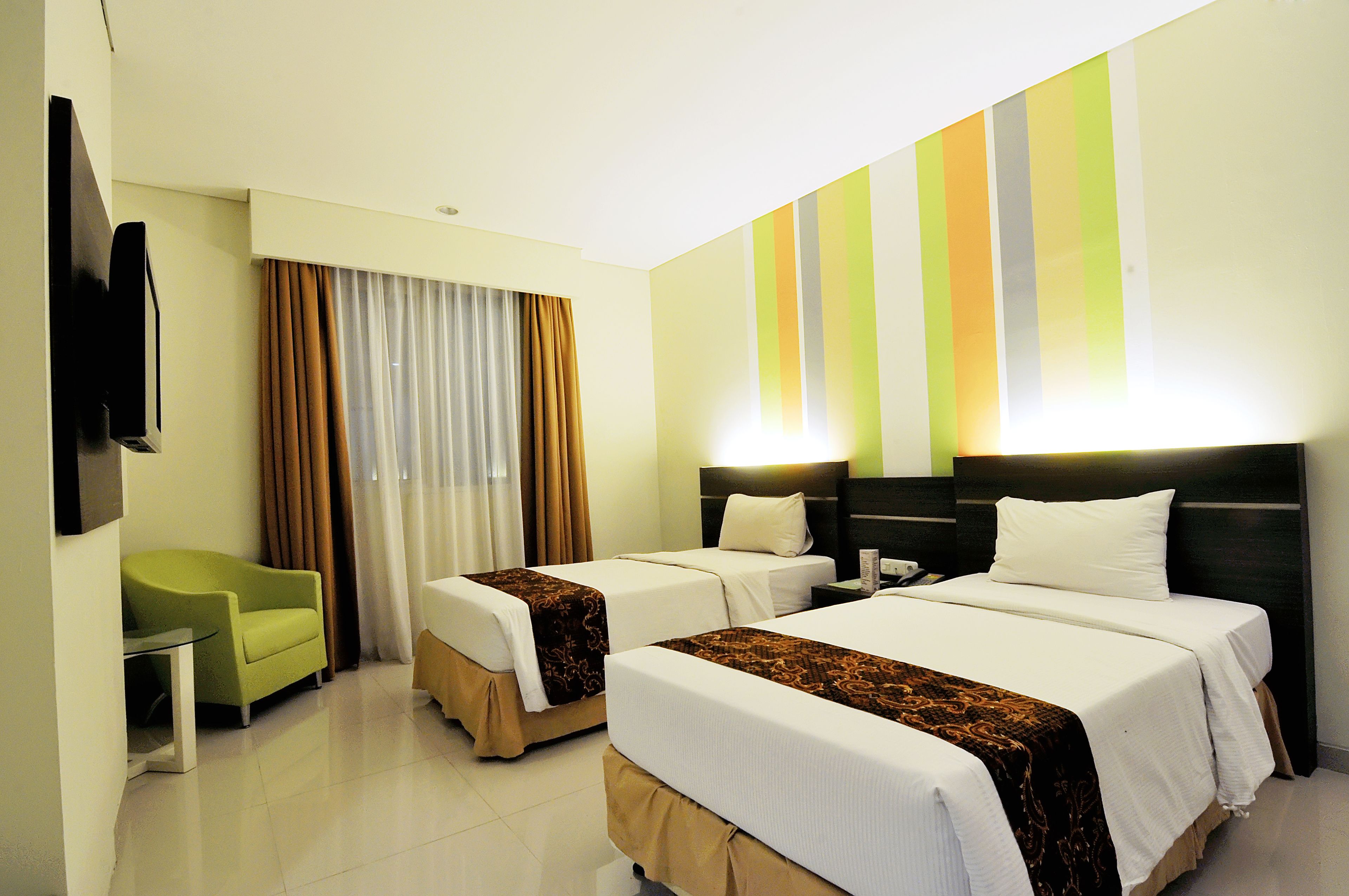A hotel room with two beds, a green armchair, a flat-screen TV, and a wall with vertical stripes.