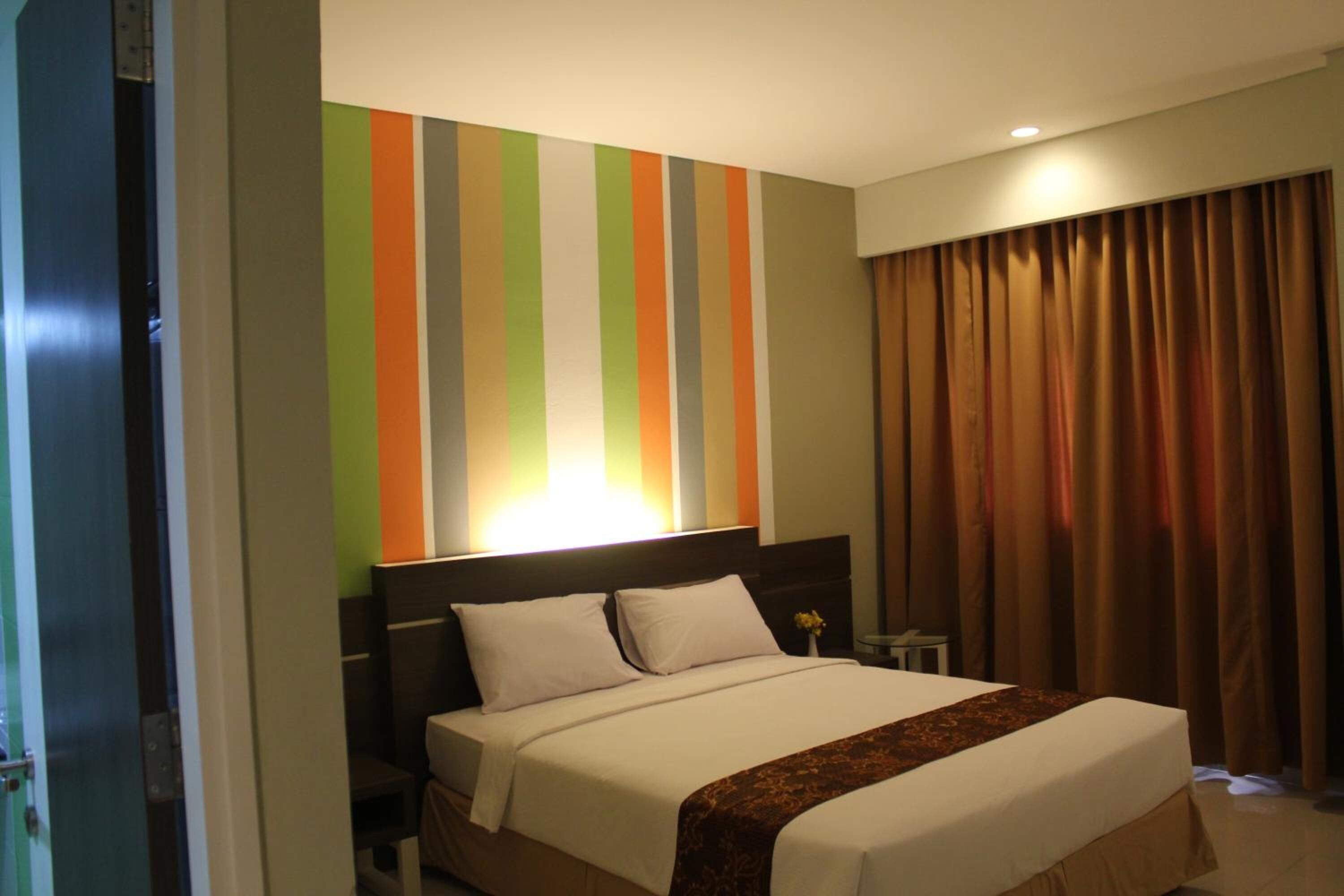 A hotel room with a large bed, a headboard with vertical stripes, a nightstand with a lamp, and a window with curtains.