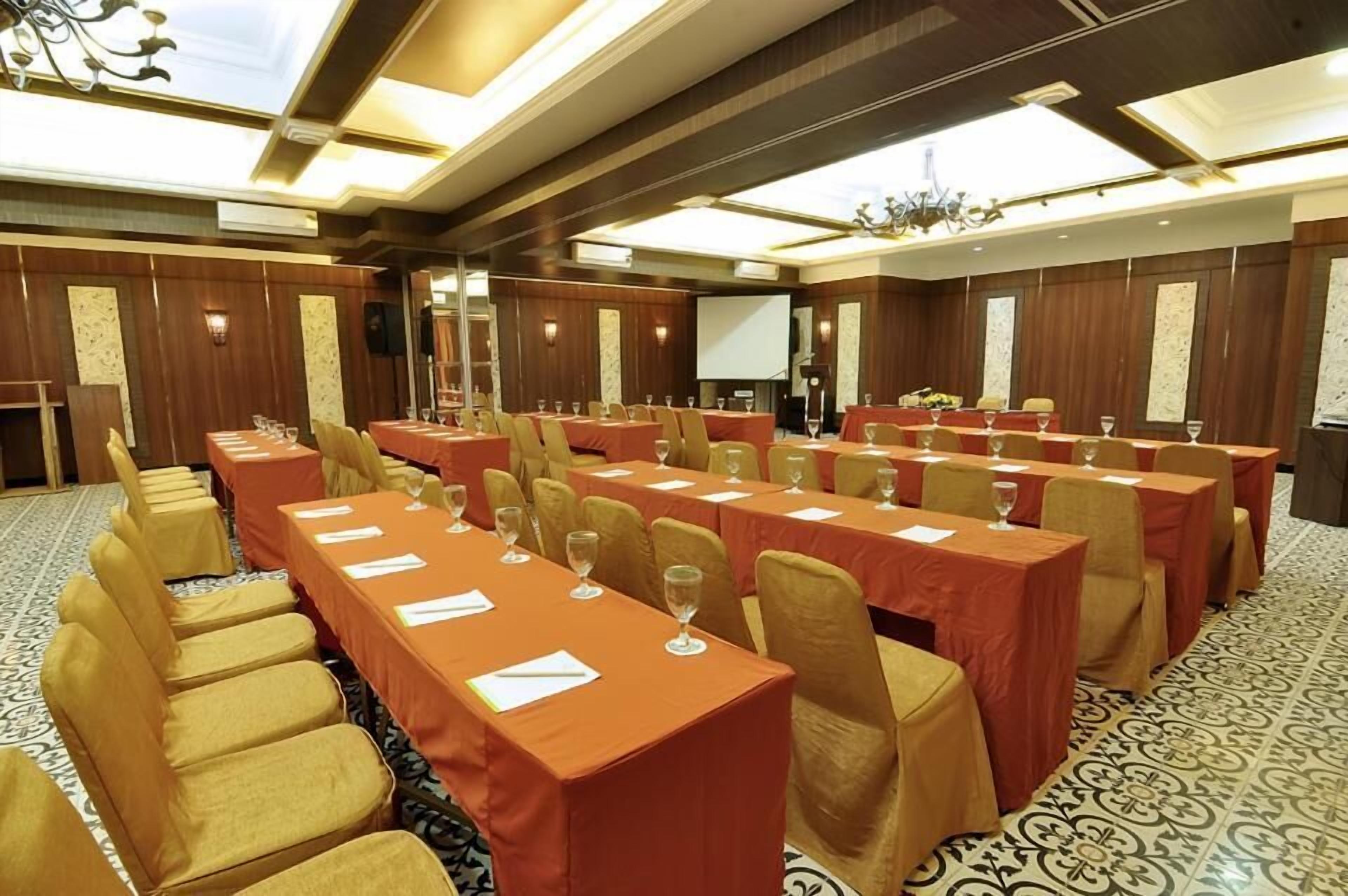 Une salle de conférence avec des rangées de tables et de chaises, équipée de projecteurs et d’écrans.