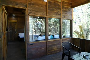 A wooden cabin with a bed, a window with a view of trees, and a table with a bowl. - Tiger N Woods Resort (Kurai)