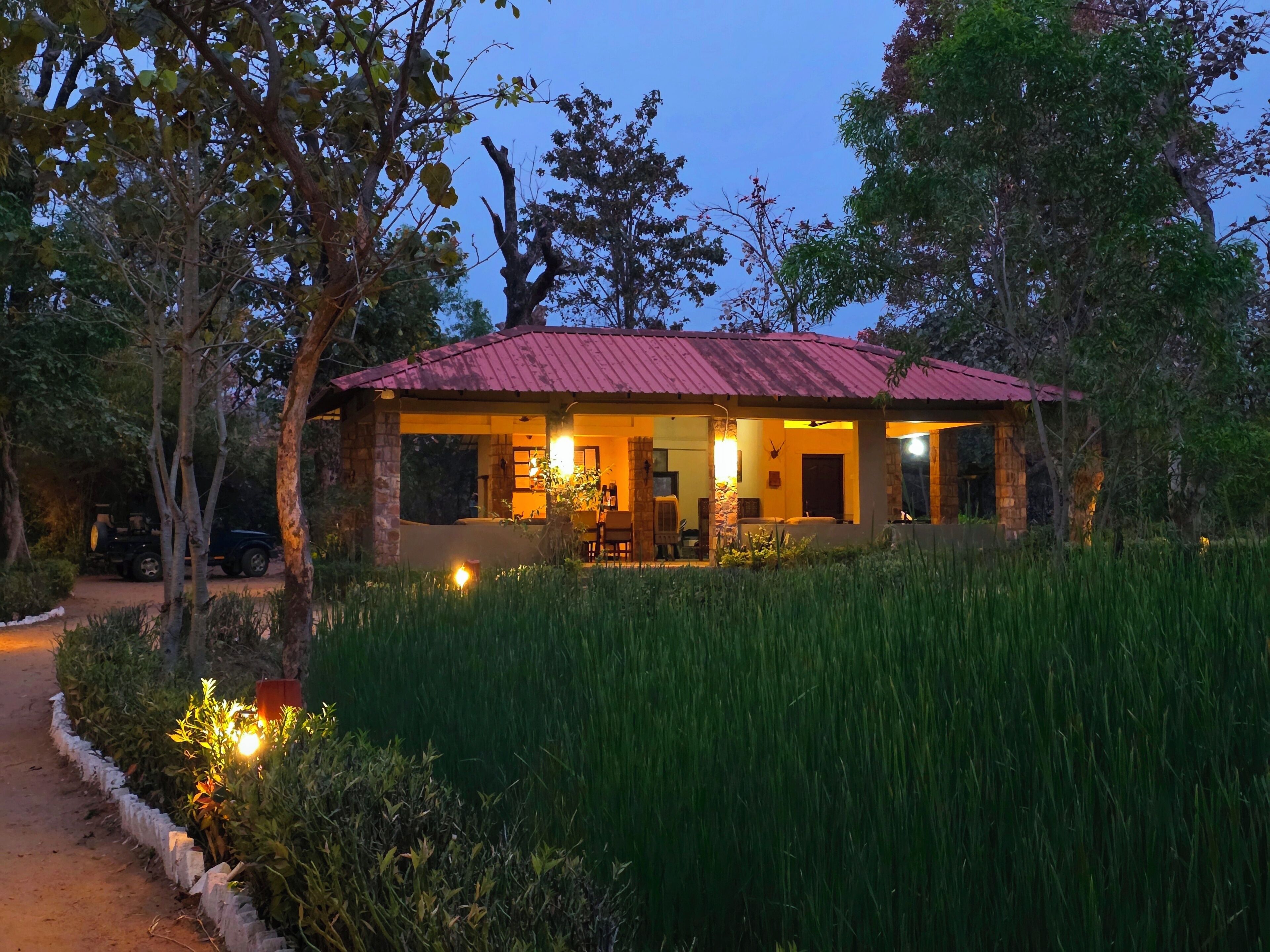 A rustic house with a red roof, surrounded by tall grass and trees, featuring a stone exterior and a visible porch area.