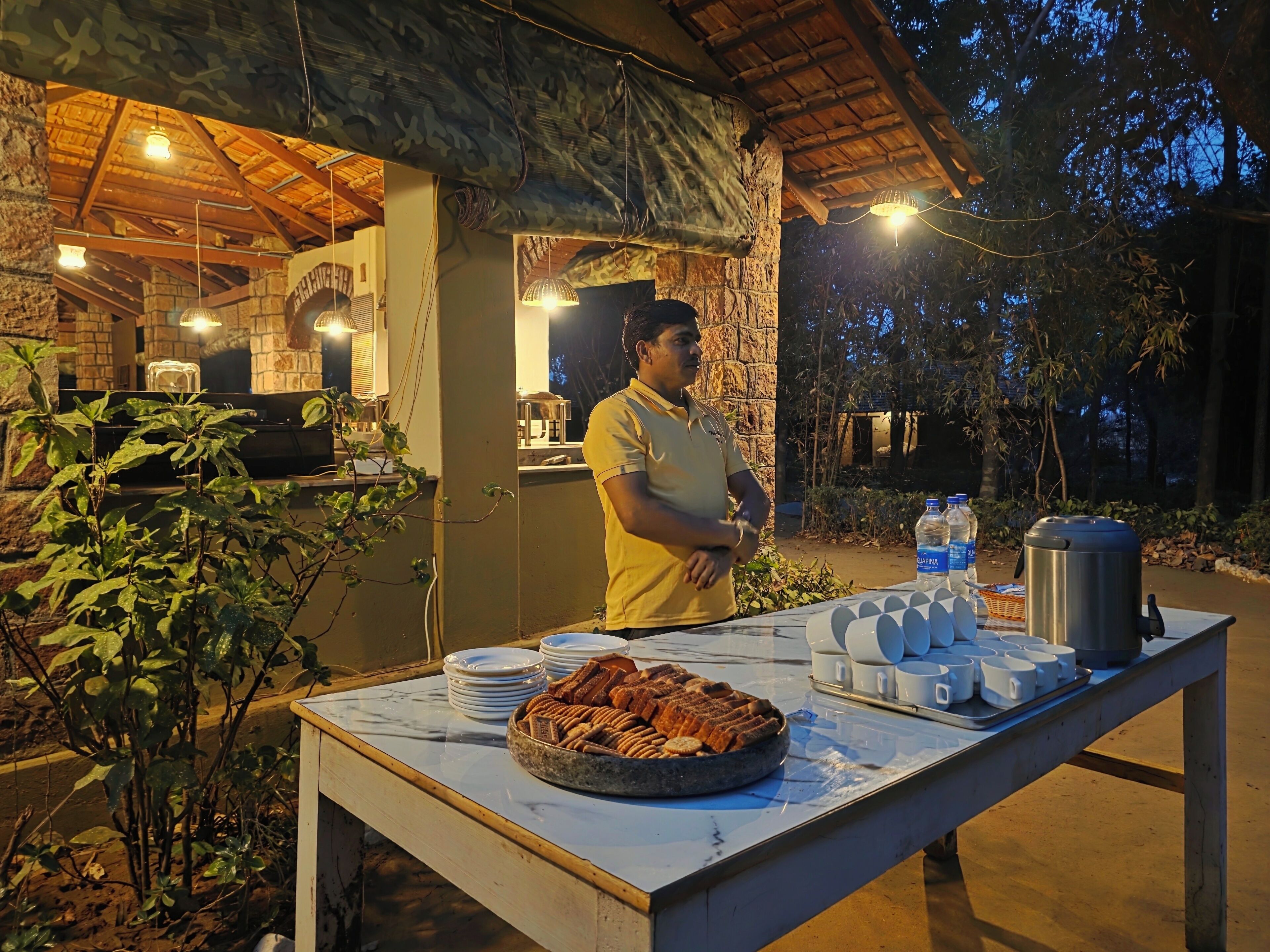 An outdoor buffet setup with a table, plates, cups, and a kettle.