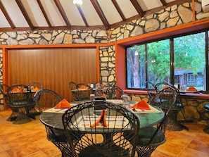 A restaurant with stone walls, wooden ceiling beams, and large windows with a view of trees. Tables are set with chairs and orange napkins. - Tiger Moon Resort (Sawai Madhopur)