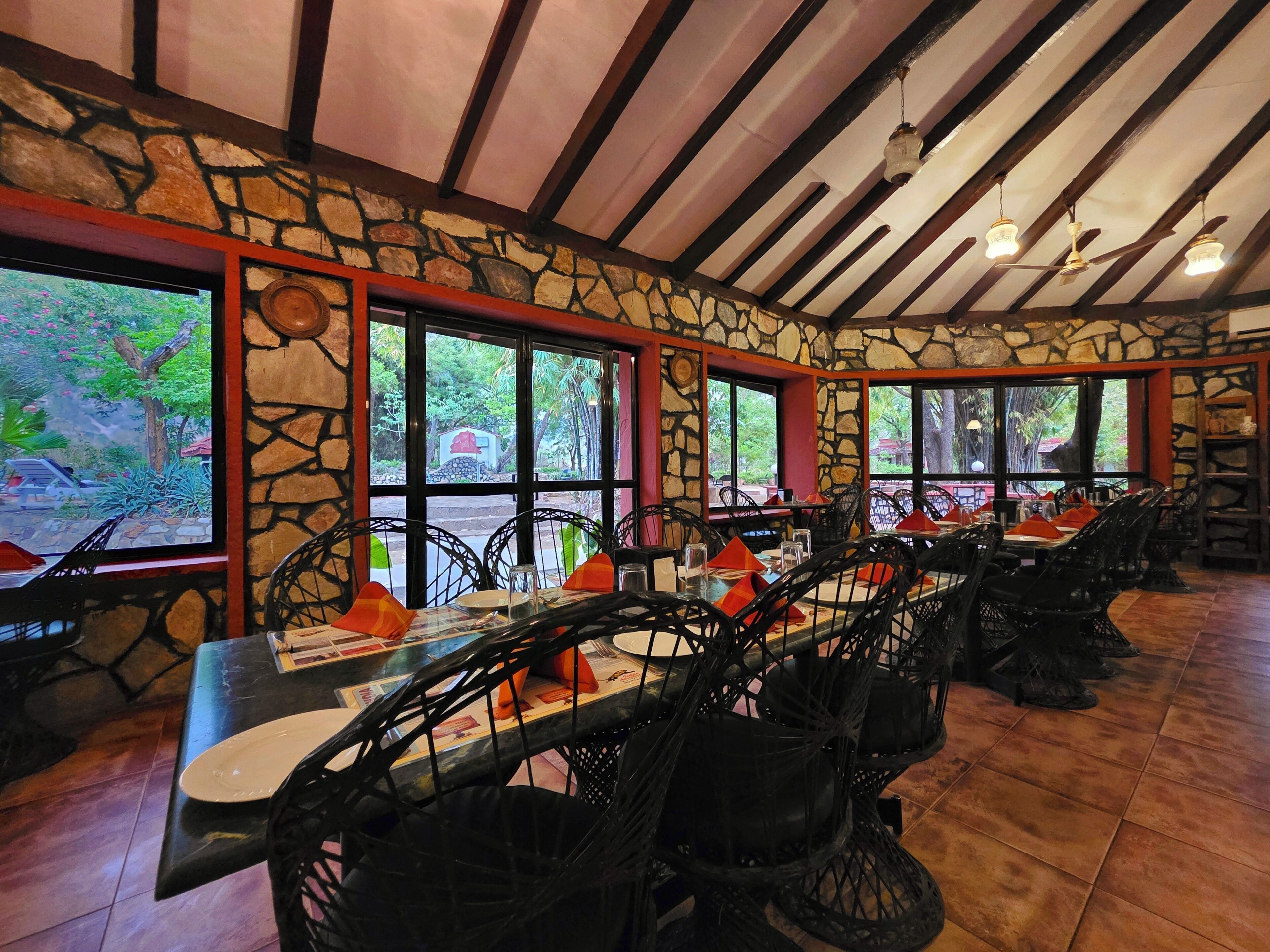 A restaurant with stone walls, wooden beams, and large windows. Tables are set with chairs and orange napkins.