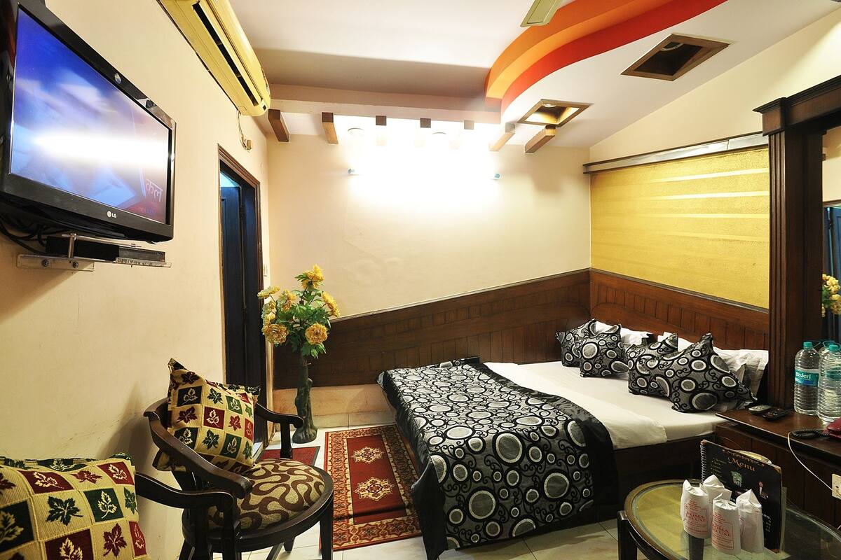 A hotel room with a bed, a TV, a chair, a small table, and a vase with flowers.