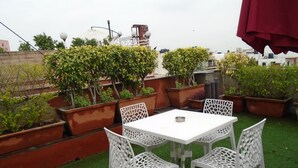 Une terrasse sur le toit, avec une table et des chaises blanches, entourée de plantes en pot et agrémentée d’un parasol rouge.