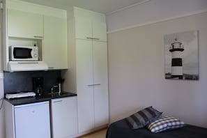 Quarto compacto com cozinha americana, uma cama com duas almofadas e um quadro de um farol na parede.
