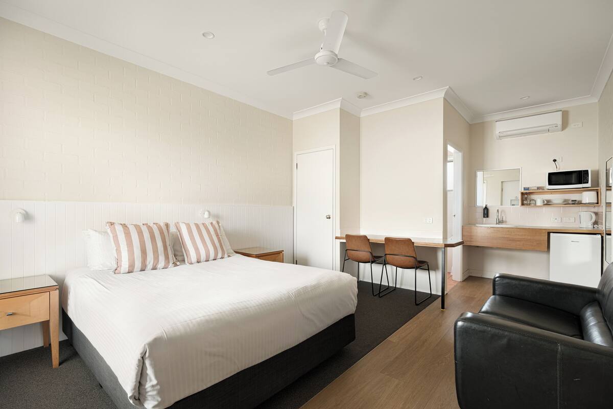 Hotelkamer met een bed, een bureau, twee stoelen, een zwarte leren bank, een plafondventilator en een magnetron.