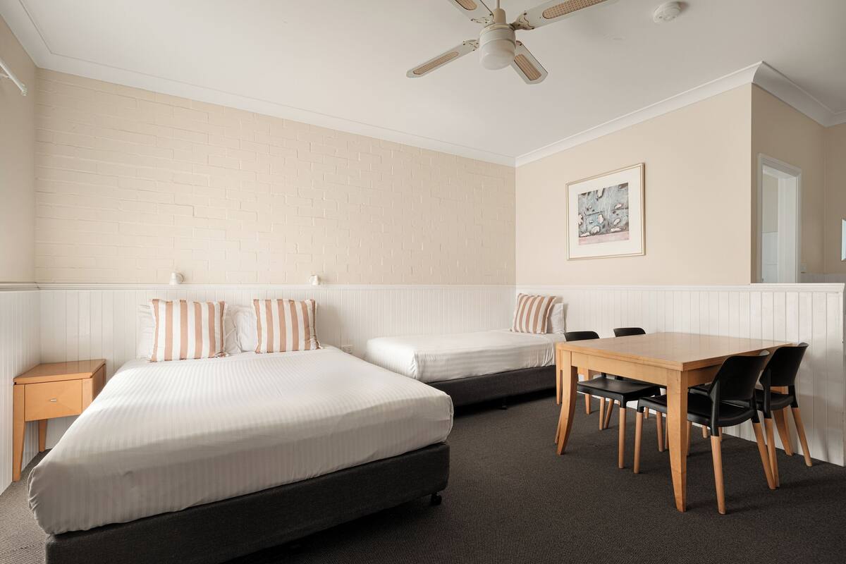 Een hotelkamer met twee bedden, een eettafel met stoelen, een plafondventilator en een inlijsting foto aan de muur.