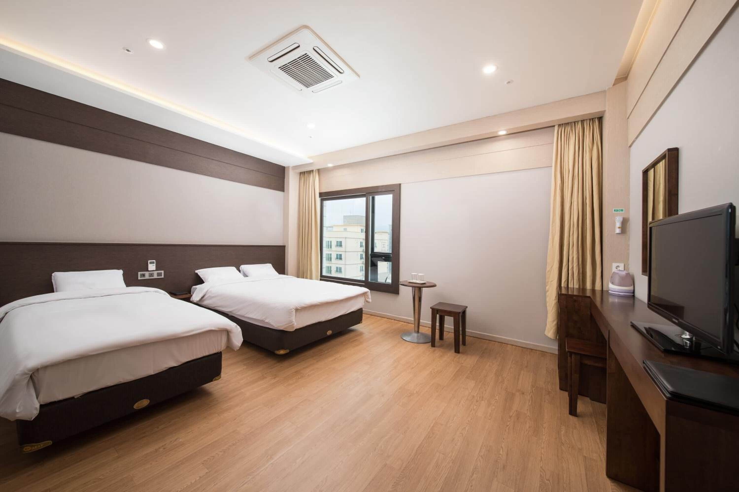 A hotel room with two beds, a TV, a small table, and a window with a view of buildings.