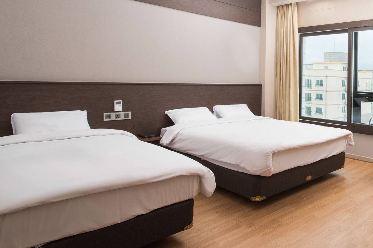 Habitación de hotel con dos camas, piso de madera, un ventanal amplio con vistas a edificios y un enchufe en la pared.