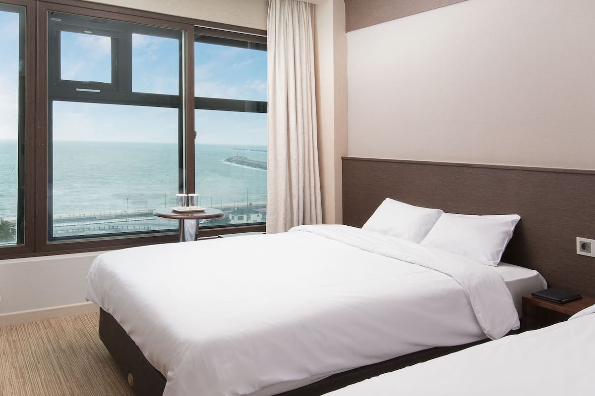 Habitación de hotel con una ventana grande que ofrece vistas al mar, una cama con sábanas blancas, una mesita de noche y un panel de control en la pared.