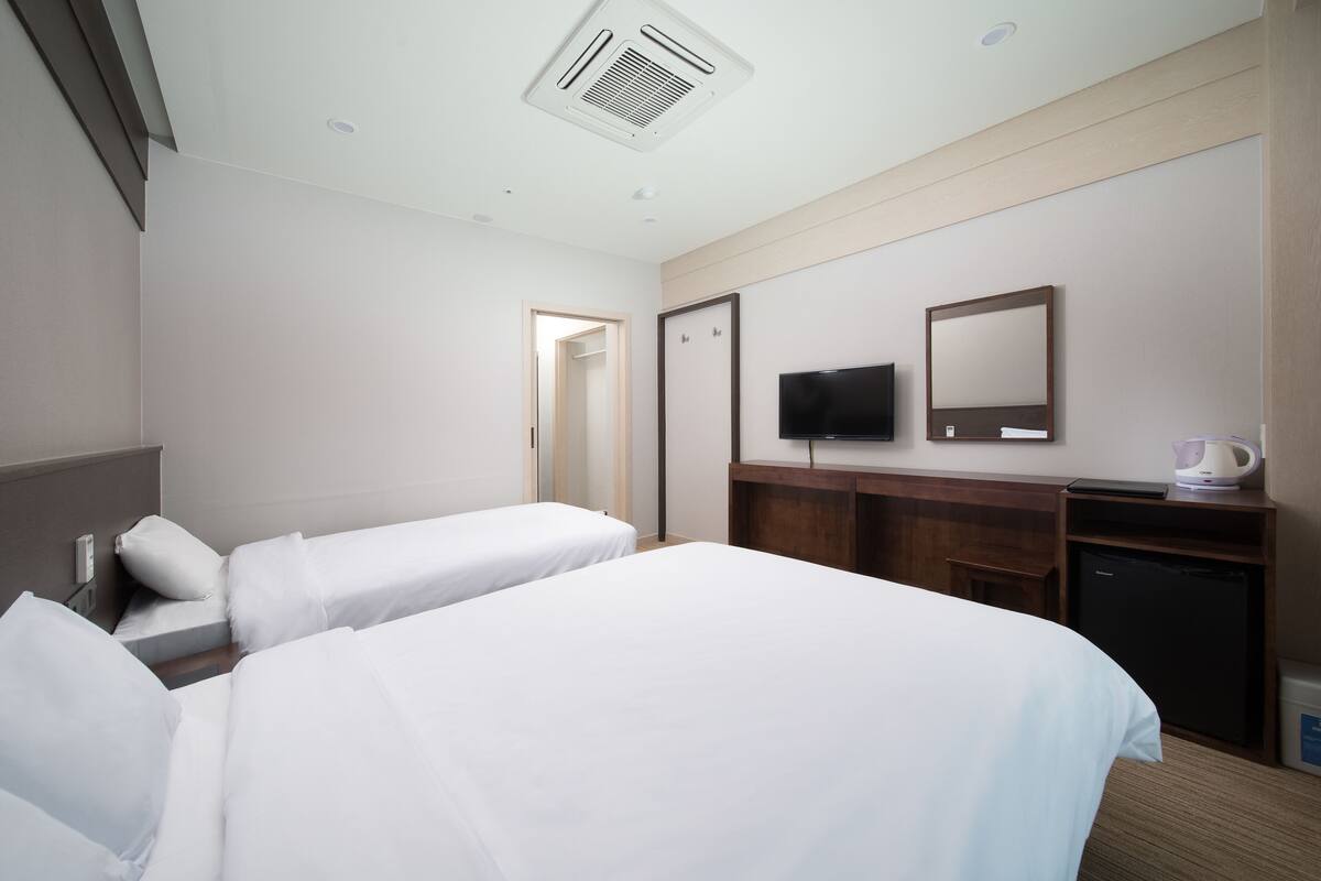 Habitación de hotel con dos camas individuales, un televisor, un espejo, un escritorio con cafetera y un armario.