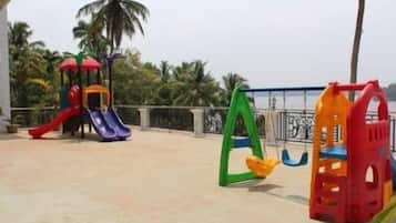Children can play safely on the outdoor play structures, including slides and swings, surrounded by a low fence and palm trees.